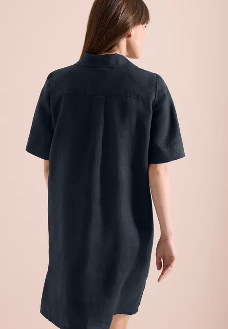 Rugaanzicht van een vrouw in een korte, donkerblauwe overhemdjurk met korte mouwen, voor een roze achtergrond.