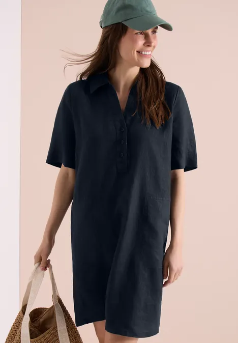 Vrouw draagt een losvallende donkerblauwe overhemdjurk, groene pet en rotan tas.