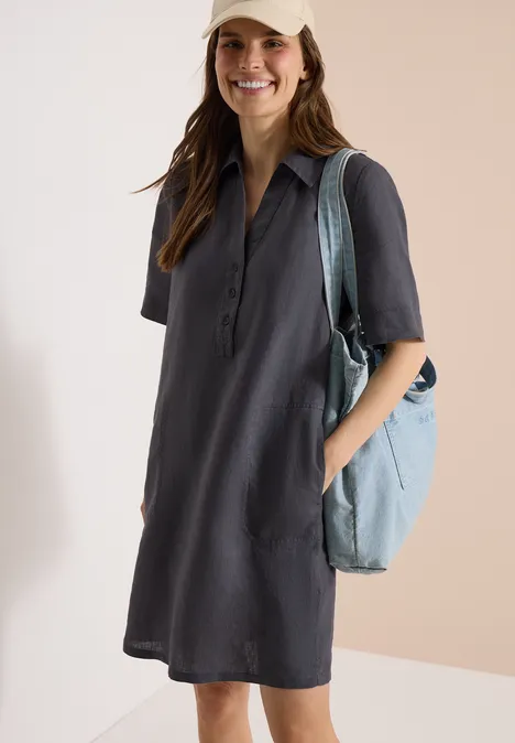 Vrouw in donkergrijze overhemdjurk met korte mouwen en lichtblauwe schoudertas, handen in de zakken.