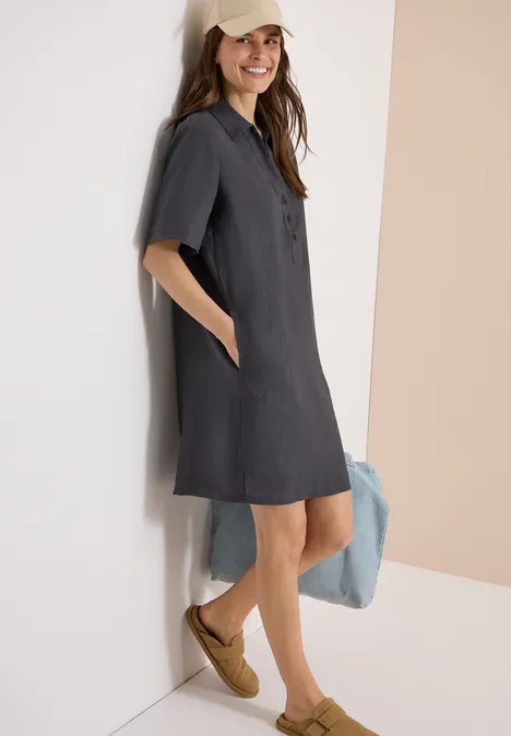 Vrouw leunt tegen een muur in een donkergrijze hemdjurk met korte mouwen en gevlochten sleehakken.