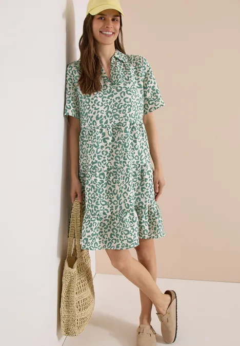 Frau in lockerem, kurzärmligem, grün-weiß geblümtem Sommerkleid mit Rüschen, trägt Basttasche und Espadrilles.