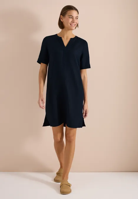 Vrouw in donkerblauwe jurk en beige instappers tegen een effen, neutrale achtergrond.