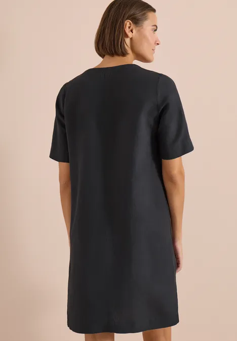 Vrouw van achteren in een eenvoudige zwarte linnen jurk met korte mouwen, gefotografeerd tegen een neutrale achtergrond.