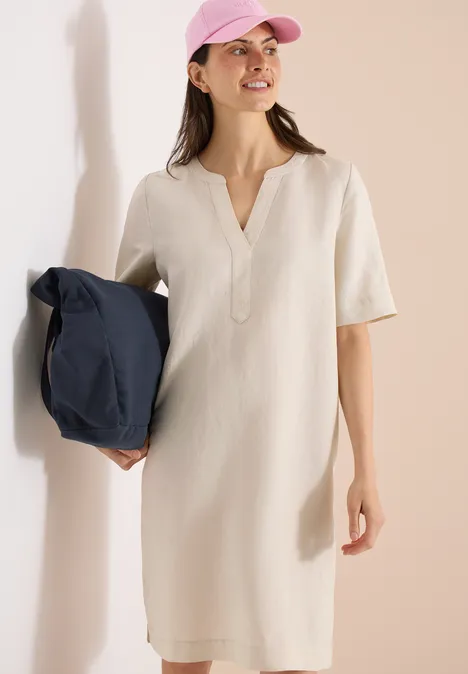 Vrouw in crème korte mouw jurk met V-hals houdt een donkerblauwe tas vast en draagt een roze pet.