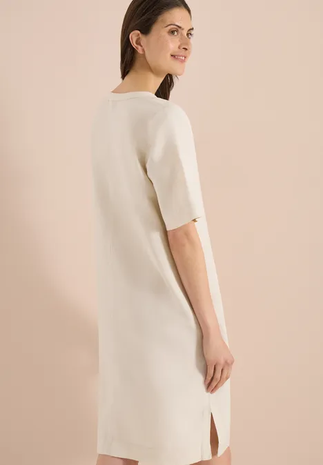 Achteraanzicht van een vrouw in een eenvoudige, crèmekleurige midi-jurk met korte mouwen en een split.