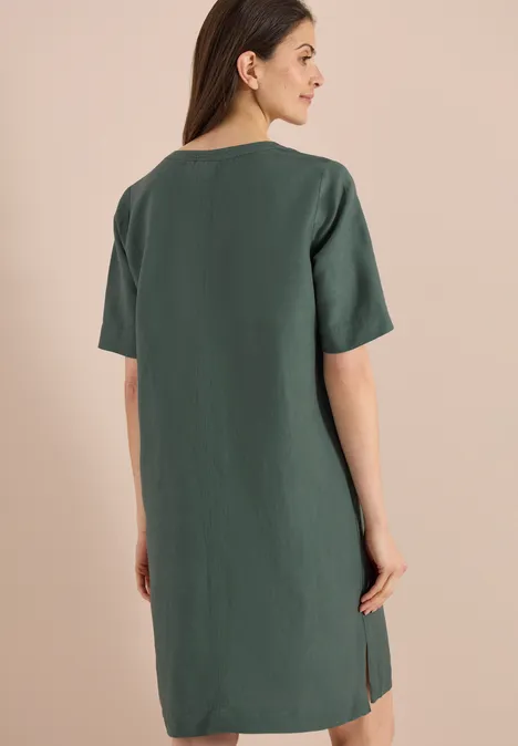 Frau von hinten in schlichten, kurzärmeligen, dunkelgrünen Kleid mit minimalem Seitenschlitz, vor neutralem Hintergrund.