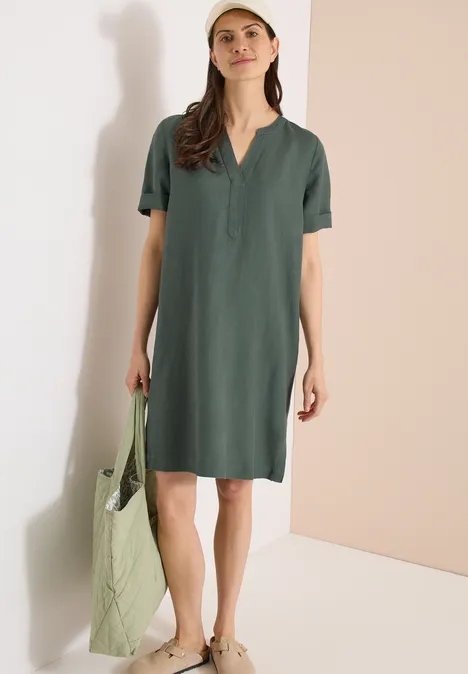 Frau in lockerem, knielangen, grünen Kleid mit V-Ausschnitt, trägt grüne Tasche und helle Slipper.