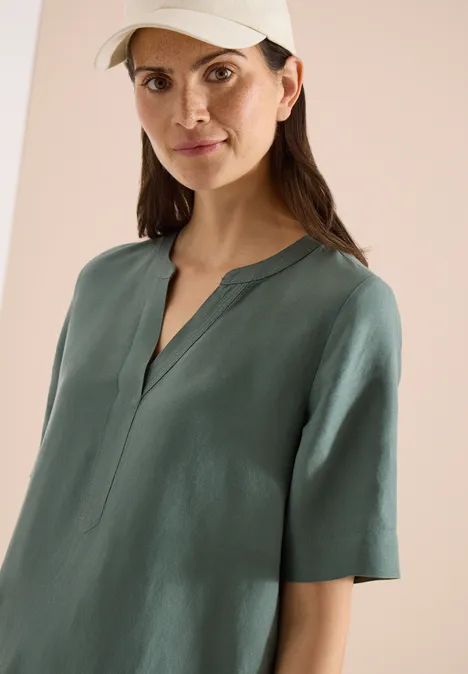 Frau mit grüner Bluse und Baseballcap, Gesicht verborgen, minimalistischer Hintergrund.