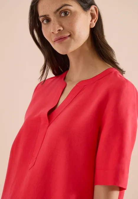 Frau mit roten Oberteil und dezentem Lächeln vor hellem Hintergrund.