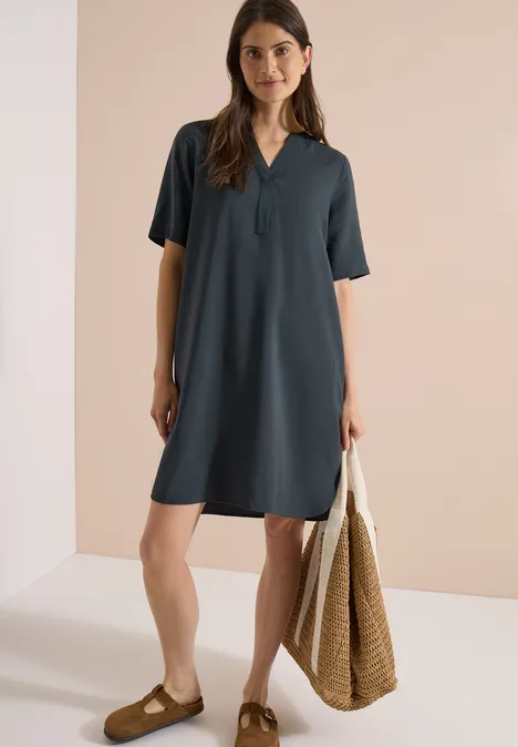 Knielanges Kleid mit Split Neck palm breeze green Frau in schlichtem, knielangem, dunkelgrauem Kurzarmkleid, mit Basttasche und flachen Schuhen.
