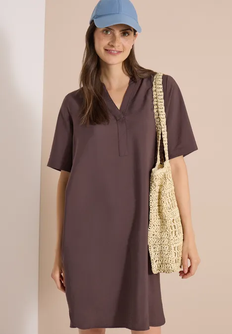 Mannequin porte une robe chemise marron à manches courtes et col en V, casquette bleue et sac en paille tressée.