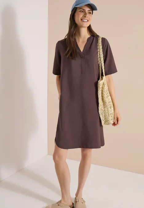 Femme en robe polo marron chocolat avec sac en paille, debout devant un fond beige.