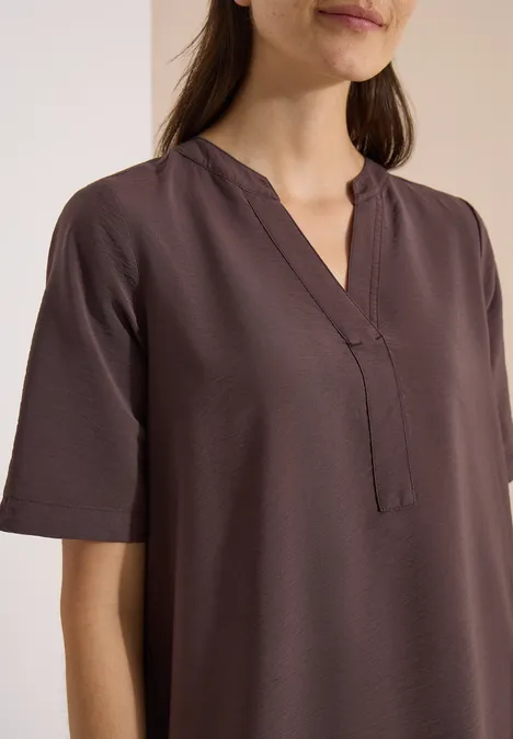 Femme portant un chemisier marron simple à manches courtes, col en V et coupe droite sur fond clair.