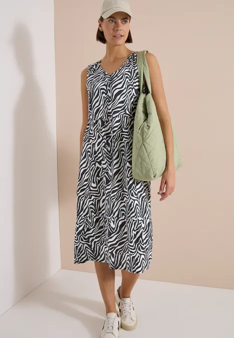 Vrouw in mouwloze midi-jurk met zebraprint, witte sneakers en grote groene tas.