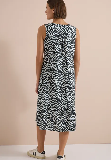 Een vrouw draagt een mouwloze midi-jurk met zwart-wit zebraprint tegen een neutrale achtergrond.