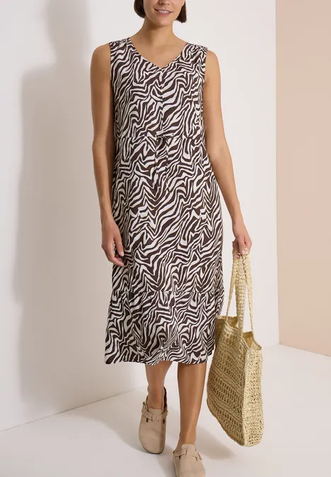 Vrouw draagt mouwloze midi-jurk met zebraprint, rotan tas en beige loafers.