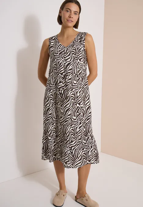 Vrouw in mouwloze midi-jurk met zebrapatroon en beige loafers voor neutrale achtergrond.