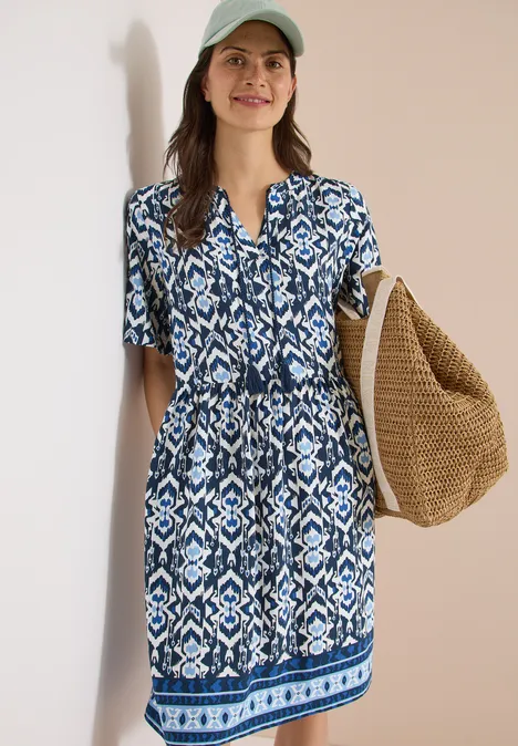 Frau trägt blau-weiß gemustertes Sommerkleid und große Strohtasche vor hellbeigem Hintergrund.