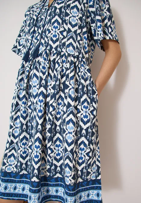Frau trägt ein langes blau-weiß gemustertes Kleid mit kurzen Ärmeln und Seitentaschen vor hellem Hintergrund.