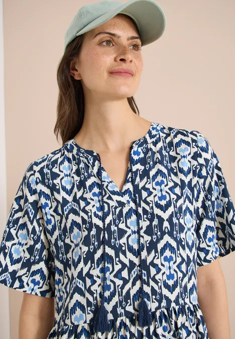 Frau trägt kurzärmeliges, blau-weiß gemustertes Top mit Quasten am Ausschnitt und grüne Baseballkappe.