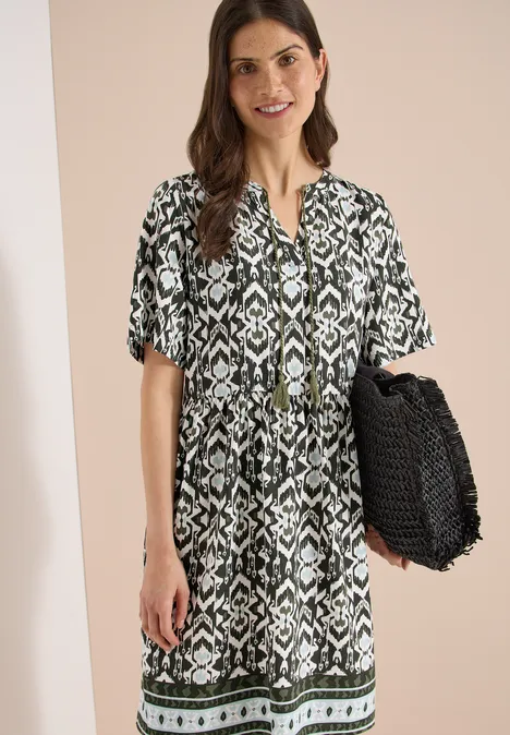 Frau in schwarz-weiß gemustertem Kurzkleid mit schwarzer Flecht-Tasche vor neutralem Hintergrund.