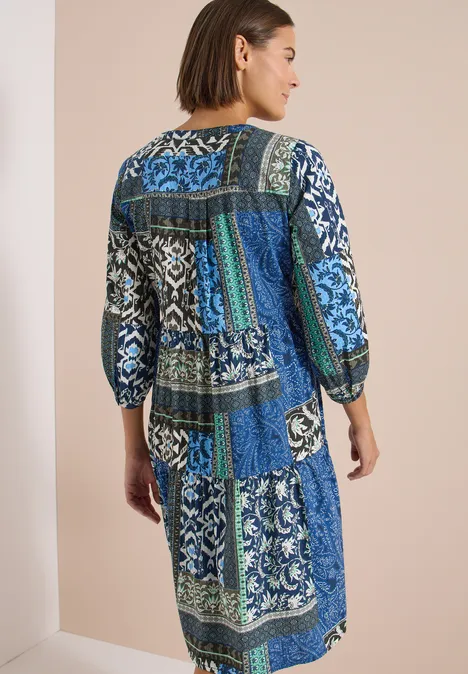 Femme vue de dos portant une robe patchwork bleu-vert aux manches 3/4 devant un mur clair.