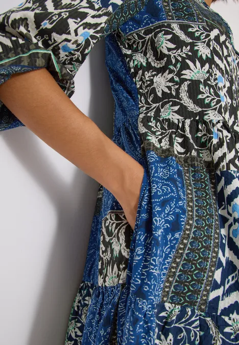 Main dans la poche latérale d'une robe longue patchwork aux motifs floraux bleus, blancs et noirs.