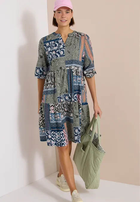 Frau in geblümtem Patchwork-Kleid mit grüner Tasche und beigen Sneakern vor hellem Hintergrund