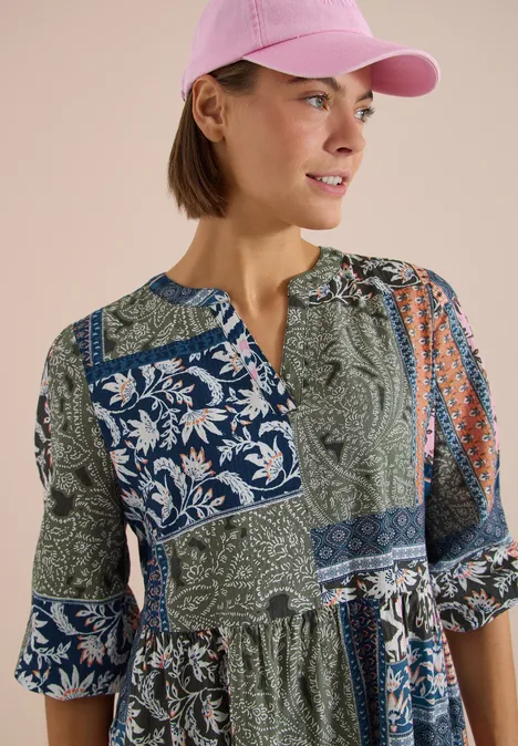 Frau mit rosa Baseballcap und gemusterter Patchwork-Bluse vor neutralem Hintergrund