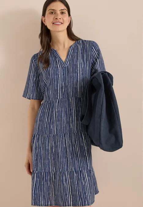 Frau in blauem, gestreiftem, knielangem Kleid mit V-Ausschnitt und halblangen Ärmeln, hält passende Jacke in der Hand.