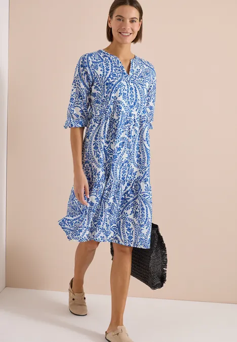 Vrouw in blauw-wit midi-jurk met paisleypatroon, V-hals, driekwartmouwen en platformsandalen.