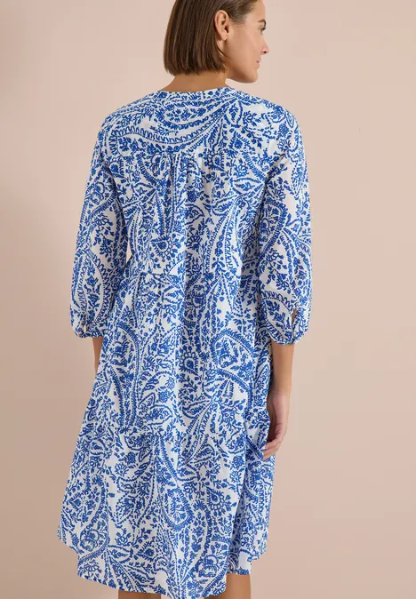 Vrouw van achteren in een losvallende, knielange jurk met blauw-wit bloemmotief voor een neutrale achtergrond.