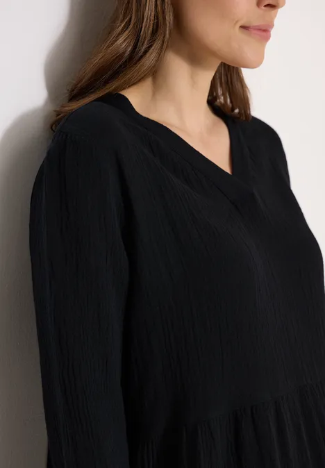 Eine Frau in Nahaufnahme trägt eine schwarze, leicht strukturierte Bluse mit V-Ausschnitt vor heller Wand.