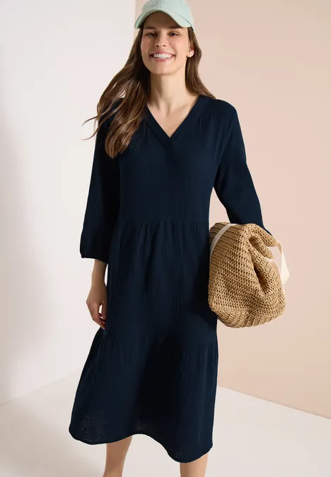 Frau in knielangem, marineblauem Kleid mit V-Ausschnitt hält Strohhut vor hellem Hintergrund.