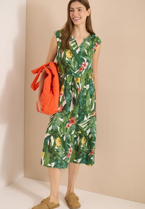 Vrouw in een groene midi-jurk met tropische bladvprint en V-hals, ze houdt een oranje gewatteerd jack vast.