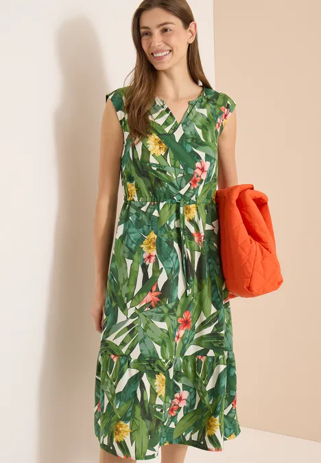 Vrouw in groene midi-jurk met tropische print en vogels, draagt een oranje doorgestikte jas.