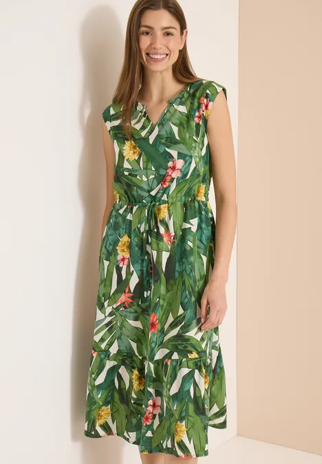 Een vrouw draagt een groene, mouwloze midi-jurk met tropische bladprint en tailleceintuur.