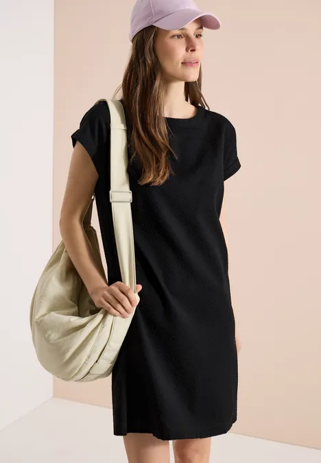 Vrouw in eenvoudig zwart jurkje, roze pet en lichtbeige schoudertas voor een rustige achtergrond.
