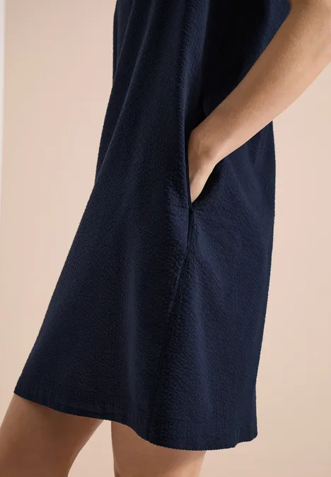 Person trägt dunkelblaues, strukturiertes Kleid mit Eingrifftasche, Hand in der Tasche vor beigem Hintergrund.