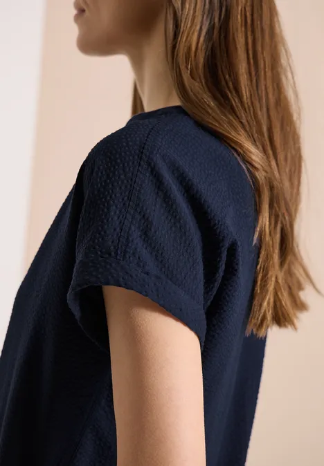 Seitliche Ansicht einer Frau in einem dunkelblauen, strukturierten T-Shirt vor neutralem Hintergrund.