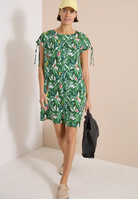 Vrouw draagt groene jurk met tropisch bladpatroon, houdt een zwarte gevlochten tas en draagt beige sandalen.