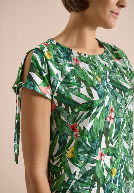 Close-up van een vrouw in een groene blouse met tropische blad- en bloemenprint, mouwen met openingen en lintjes.