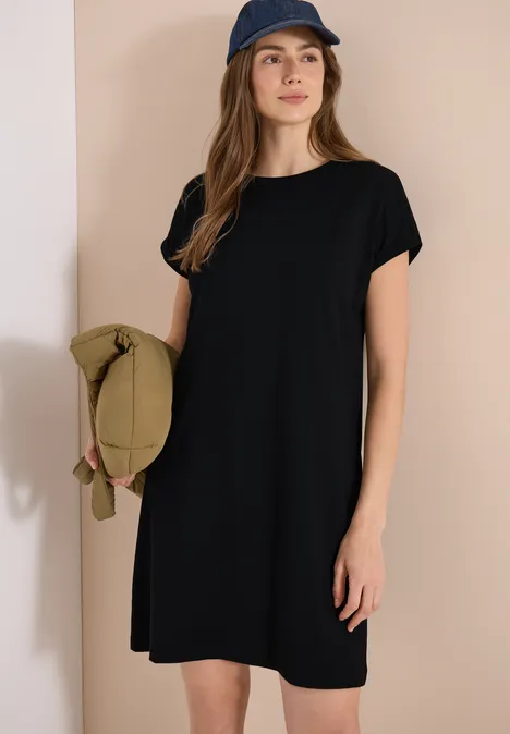 Vrouw in zwarte T-shirtjurk en pet, met een groene tas voor een beige muur.