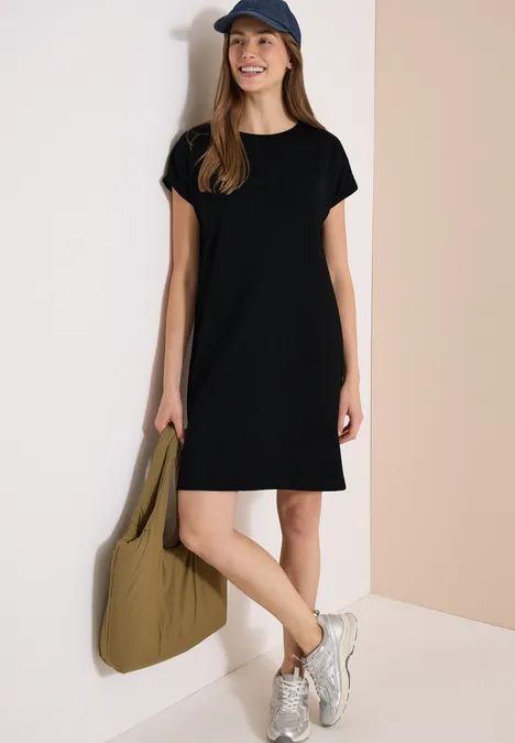 Vrouw in zwarte T-shirtjurk, pet, witte sneakers en grote olijfgroene tas.