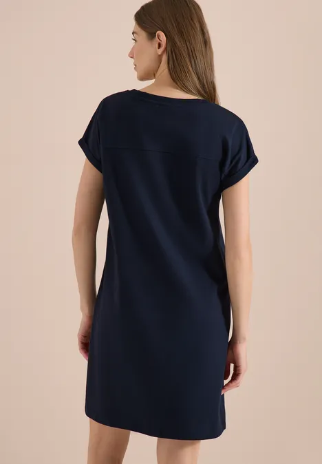 Vrouw van achteren in eenvoudig donkerblauw t-shirtjurk tot op de knie voor beige achtergrond
