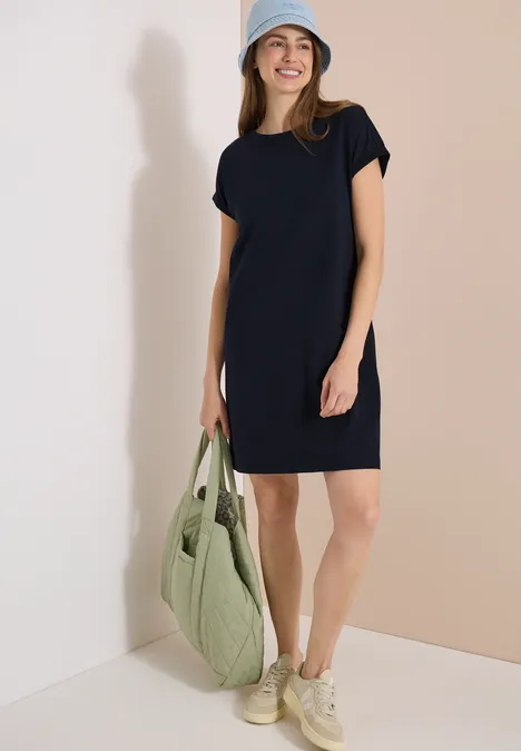 Vrouw in losvallende donkerblauwe t-shirtjurk, lichte sneakers, pet en grote groene gewatteerde tas.