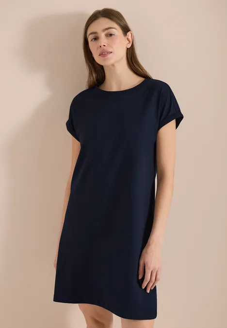 Vrouw draagt een eenvoudig donkerblauw T-shirtjurk met korte mouwen tegen neutrale achtergrond.