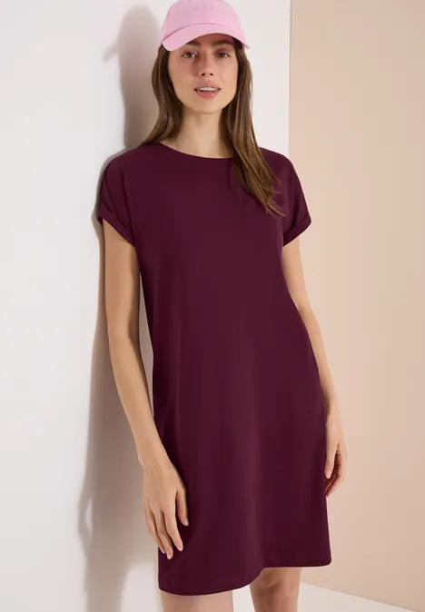 Vrouw leunt tegen een lichte muur, draagt een losse T-shirtjurk met korte mouwen in bordeaux.