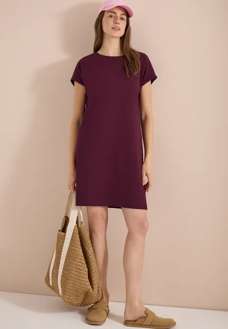 Vrouw in losvallende paarse jurk met strozak en platte muiltjes, voor een effen beige achtergrond.
