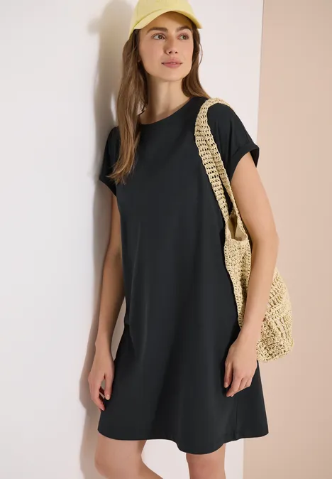 Een vrouw draagt een zwarte T-shirtjurk en houdt een gevlochten strooien tas vast tegen een lichte achtergrond.